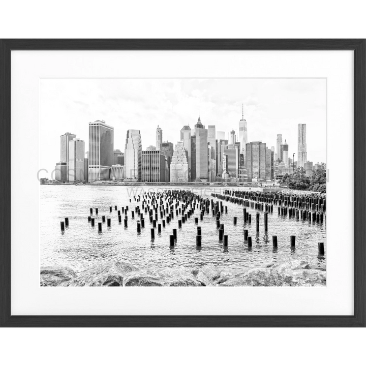 Schwarz-weiß-Fotografie von Holzpfählen im Wasser mit Manhattans Skyline im Hintergrund