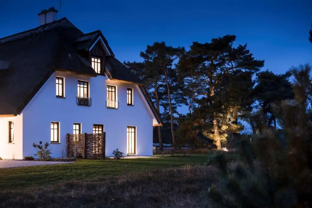 Weißes Häuschen im Landhausstil mit beleuchteten Fenstern in der Dämmerung.